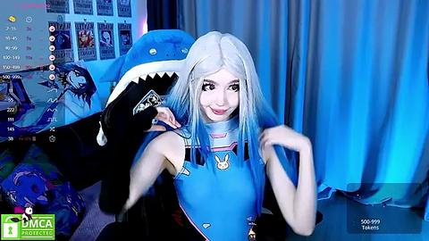 Media: Video of a young woman with pale skin, platinum blonde hair, and a blue cosplay outfit, posing playfully in front of a shark head prop, surrounded by gaming screens and blue curtains.