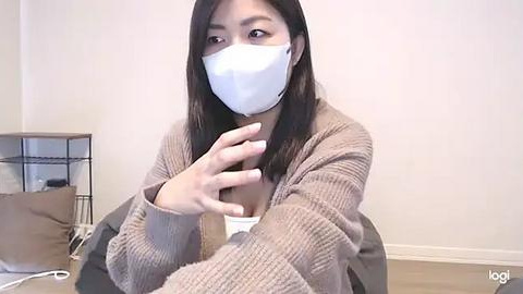 Media: Video of a young Asian woman with long black hair, wearing a white face mask, beige cardigan, and a white top, sitting indoors with a white wall and wooden floor, holding her chest.