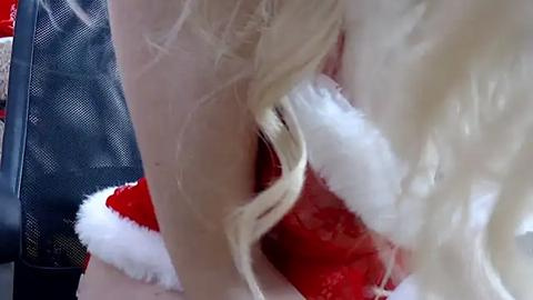 Media: Video of a blonde woman with long hair in a red and white Santa hat, sitting on a black mesh chair, viewed from the side, highlighting her hair and festive attire.