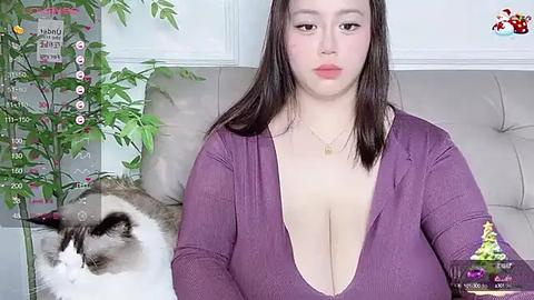 Media: A video of a plus-sized woman with light skin and long dark hair, wearing a low-cut purple top, sitting next to a fluffy white cat. The background features a green plant and a wall calendar.