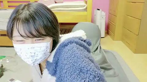 Media: Video of an Asian woman in a blue plush bear outfit with a white mask, sitting on a gray cushion on a light yellow carpeted floor. The background shows a yellow crib with pink sheets, a wooden dresser, and a white air purifier.