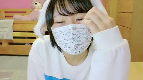 Media: Video of an Asian girl with straight black hair, wearing a white mask with cartoon animals, a white hoodie, and white bunny ears, seated in a pink room with a wooden bed and stuffed toys.