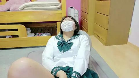 Media: Video of a young woman with pale skin and short brown hair wearing a white school uniform with a green bow, a face mask, and sitting on a light wooden floor, surrounded by a bunk bed and yellow dresser.