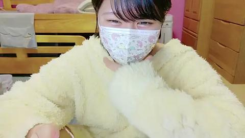 Media: Video of an Asian woman with short black hair, wearing a white face mask and a fluffy yellow bathrobe, sitting in a cozy, wooden bed and dresser room.