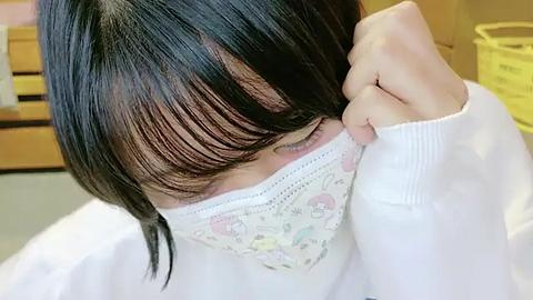 Media: Video of an Asian woman with short, straight black hair, partially covering her face with a floral-patterned mask, wearing a white sweater. Background features a wooden cabinet and yellow plastic bins.