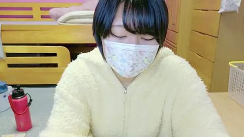 Media: Video of an Asian woman with short black hair, wearing a white mask and yellow fuzzy jacket, sitting in a brightly lit bedroom with a wooden bed and yellow dresser.