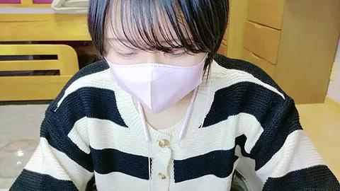 Media: Video of an East Asian woman with short black hair, wearing a white face mask and a black-and-white striped sweater, sitting in a yellow and white room with wooden furniture.