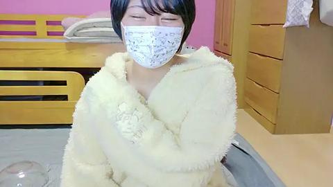 Media: Video of an East Asian person in a fluffy yellow bathrobe, wearing a white surgical mask, seated on a bed with wooden furniture and pink walls.