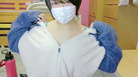 Media: Video of an Asian woman in a blue and white fuzzy cardigan, white mask, and black hair, standing in a bedroom with yellow furniture.