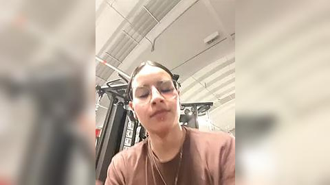 Media: A blurry video of a woman with light skin and dark hair in a brown T-shirt, using a gym machine in a gym with white walls and ceiling.