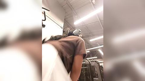 Media: Video of a person in a dark shirt, leaning forward, with a blurred background featuring gym equipment and fluorescent lighting.