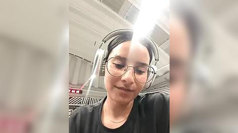 Media: Video of a young woman with fair skin, dark hair, and glasses, wearing headphones, taking a selfie in a dimly lit airport lounge with white and red signage.