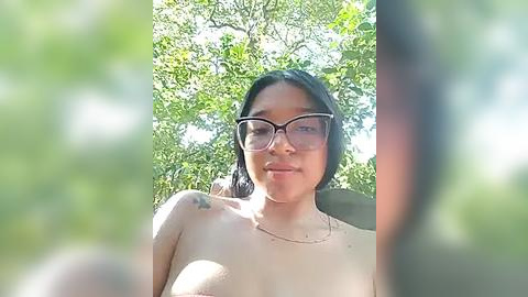 Media: Video of a smiling Asian woman with glasses and shoulder-length black hair, wearing a sheer top, against a backdrop of green trees, suggesting a sunny, outdoor setting.