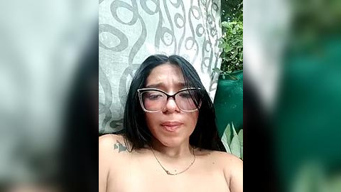 Media: A video of a topless, light-skinned Asian woman with long black hair, glasses, and a tattoo on her shoulder, standing in front of a leafy green backdrop.