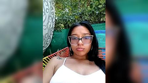 Media: Video of a young woman with medium brown skin, long black hair, and glasses, wearing a white tank top, sitting on a colorful chair surrounded by greenery.