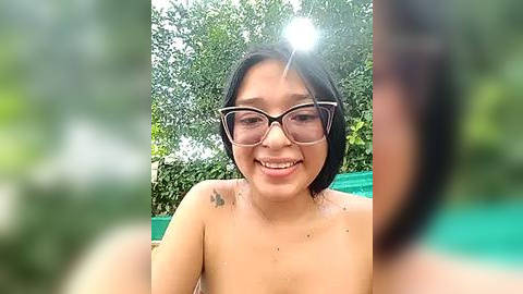 Media: Video of a smiling Asian woman with shoulder-length black hair, wearing large glasses, topless, with a tattoo on her right shoulder, against a blurred green background.