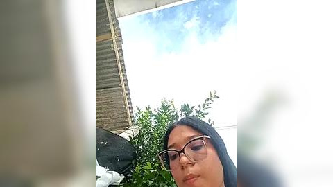 Media: Video of a woman with glasses and a black hijab, taken from a low angle. She is indoors with a bright blue sky and green foliage visible in the background.