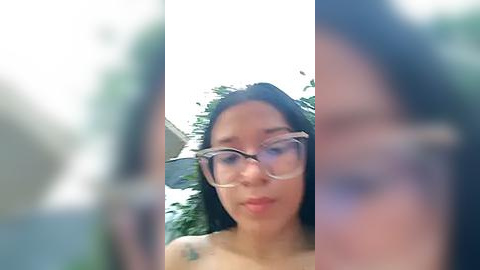 Media: A blurry video of a woman with glasses, dark hair, and a tattoo on her shoulder, standing in a garden with greenery.