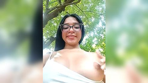 Media: Video of a smiling, light-skinned woman with glasses, wearing a white sleeveless top, outdoors amidst green foliage. Her medium-sized breasts are partially exposed.