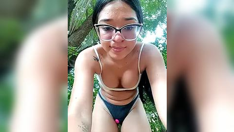 Media: A video of a young Asian woman with glasses and dark hair, wearing a revealing blue and beige bikini, leaning forward with a playful expression, surrounded by lush greenery.