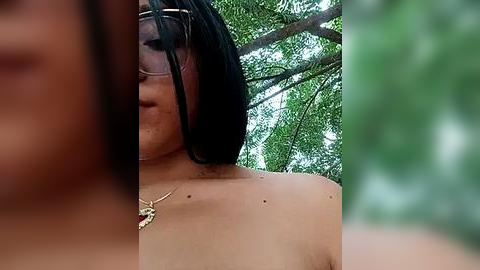 Media: Video of a close-up, partially obscured, topless Asian woman with short black hair and glasses, wearing a gold necklace, set against a green leafy background.