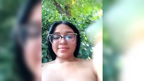 Media: A video of a topless woman with medium brown skin, wearing glasses, smiling, standing in a lush, green forest. Her long black hair falls over her shoulders.