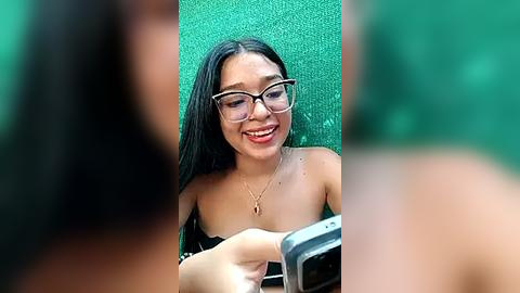 Media: A video of a smiling, young Asian woman with long black hair and glasses, seated on a green mat, taking a selfie.