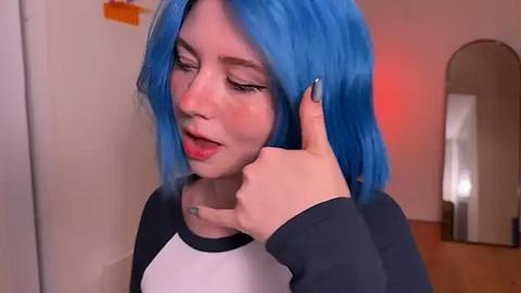 Media: Video of a young woman with bright blue hair, fair skin, and a black and white long-sleeve shirt, giving a thumbs-up while holding her ear, in a simple, beige-walled room.