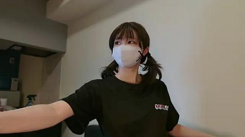 Media: Video of a young woman with long brown hair, wearing a black t-shirt and white face mask, standing indoors with beige walls and a dark door in the background.