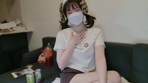 Media: Video of a young Asian woman with pale skin, wearing a white T-shirt, brown shorts, and leopard-print headphones, seated on a gray couch, covering her mouth with her hand, amid scattered food and drinks.