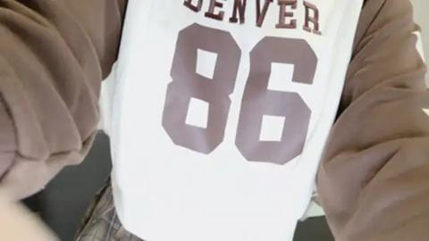 Media: Video of a person wearing a white t-shirt with large, bold brown numbers \"86\" and the word \"DENVER\" in red. The person is standing with arms outstretched, wearing a brown jacket.