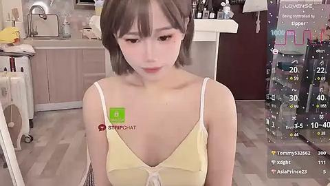 Media: Video of an East Asian woman with fair skin, short brown hair, wearing a yellow spaghetti-strap top, sitting in a modern kitchen. Background includes cabinets, a sink, and a colorful digital display.