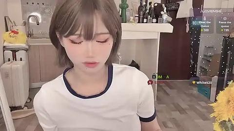 Media: Video of a young Asian woman with short, straight brown hair, wearing a white t-shirt, sitting in a kitchen. Background shows kitchen appliances, bottles, and a \"Loverly\" display.