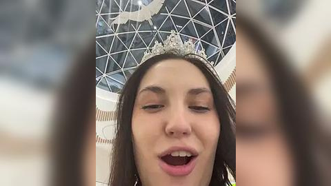 Media: Video of a young woman with long dark hair, wearing a sparkling tiara, singing with her mouth open, against a blurred, modern architectural backdrop with a large glass dome.