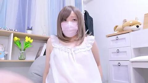 Media: Video of an East Asian woman with shoulder-length brown hair, wearing a white mask, white dress, and seated in a bedroom with white walls, yellow flowers, and a teddy bear.