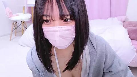 Media: Video of an Asian woman with long black hair, wearing a pink surgical mask, light blue cardigan, and a low-cut white top, sitting in a pink room with white bedding and a white chair.