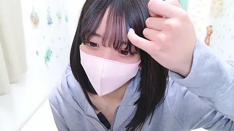 Media: A video of an Asian woman with straight black hair and a pale pink face mask, adjusting her hair in a bathroom with pastel-colored tiles. She wears a light gray hoodie.