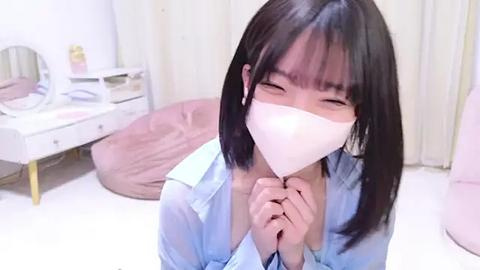 Media: Video of an East Asian woman with straight black hair, wearing a light blue shirt and a white mask, kneeling on a white floor, in a softly lit room with beige furniture.