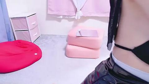 Media: Video of a minimalist room with a red beanbag, pastel pink ottoman, and a black leather jacket draped on a white wall.