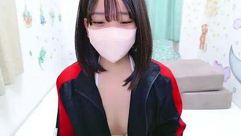 Media: Video of an Asian woman with straight black hair, wearing a white mask and black-and-red jacket, standing in a clinical setting with white walls, medical equipment, and a bed.