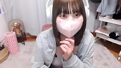 Media: Video of an East Asian woman with straight, shoulder-length brown hair, wearing white headphones, a light blue jacket, and a pink face mask. She is kneeling on a light carpet in a cluttered room with a pink storage bin, a white shelf, and a lamp.
