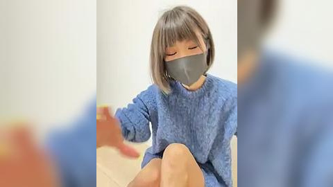 Media: Video of an Asian woman with short gray hair, wearing a blue sweater, gray face mask, and seated on a wooden floor. Background shows blurred figures in blue.