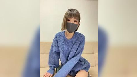 Media: Video of a young Asian woman with straight, shoulder-length brown hair, wearing a blue, textured sweater, black face mask, and sitting on a beige couch against a plain white wall.