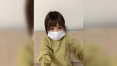 Media: A video of a young Asian girl with short brown hair, wearing a white surgical mask, seated on a beige couch, indoors, with a blurred background.