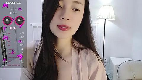 Media: Video of a young Asian woman with long black hair, wearing a pink shirt, in a modern, well-lit room with white furniture and a lamp.