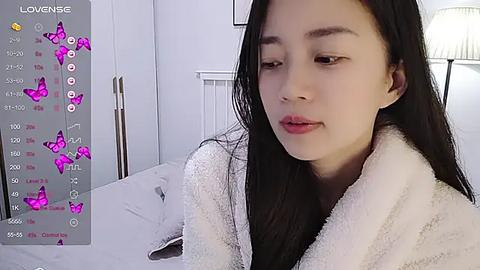 Media: Video of a young East Asian woman with long black hair, light skin, and slender build, wearing a white fluffy bathrobe, looking contemplative in a modern, minimalistic white bedroom.