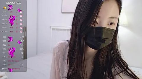 Media: A video of a young Asian woman with long black hair, wearing a black face mask and a light-colored top, indoors, in a modern, minimalist room with white walls and a bed.