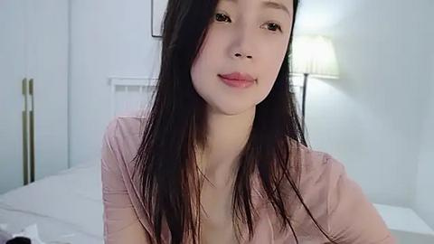 Media: A video of a young Asian woman with long black hair, fair skin, and small breasts, wearing a light pink shirt, smiling softly. She's indoors, seated on a bed with white sheets, next to a bedside lamp.