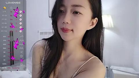 Media: A video of an East Asian woman with long black hair, fair skin, and a slim physique, wearing a beige tank top, smiling in a modern, softly lit bedroom.