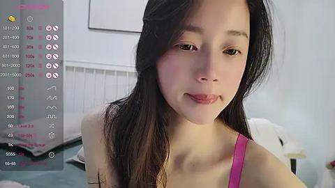 Media: A video of a young Asian woman with long black hair, wearing a pink top, smiling in a bedroom with white walls and a bed.